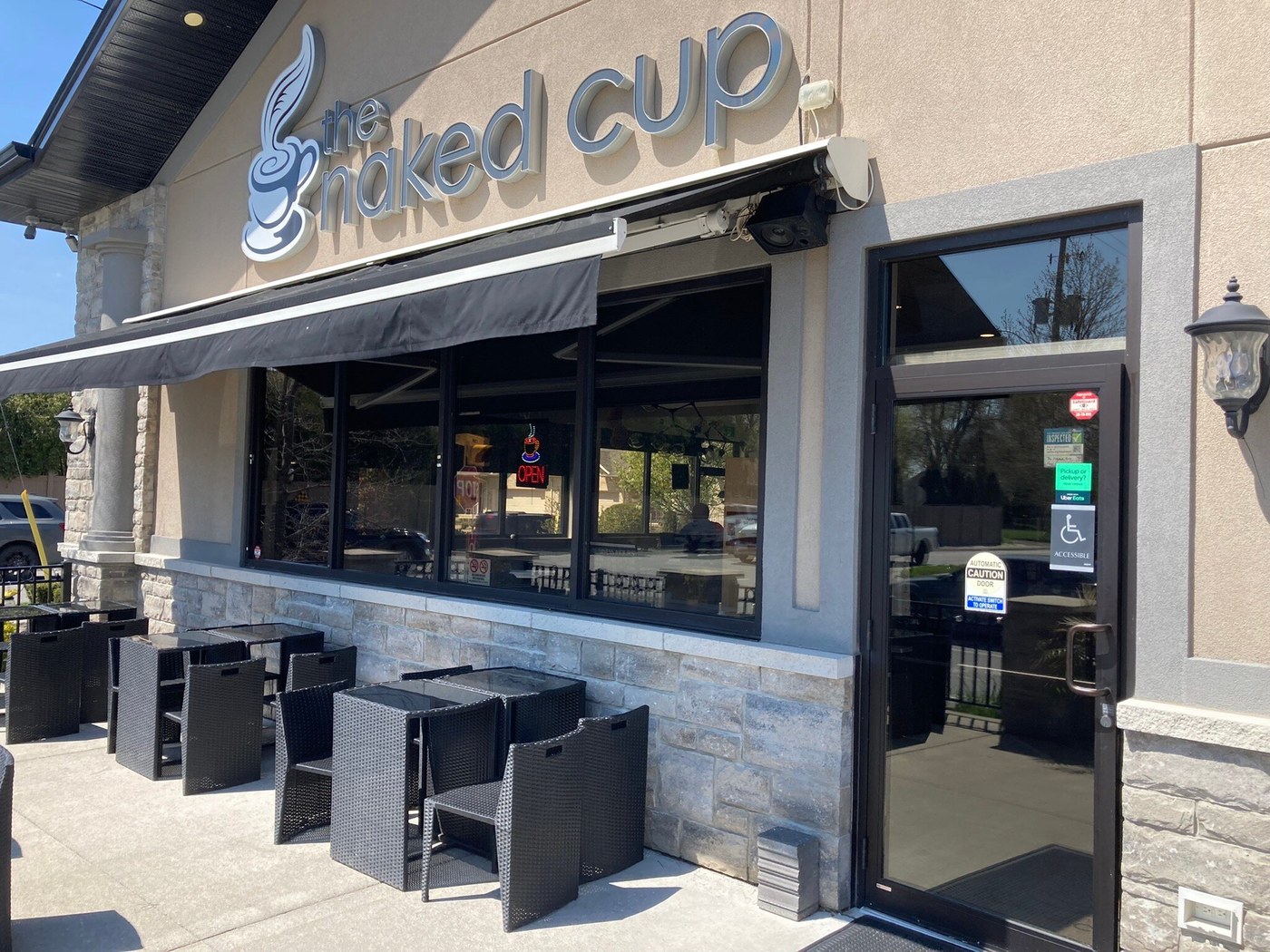 The Naked Cup storefront with raised lettering, a black awning and pavement tables and chairs